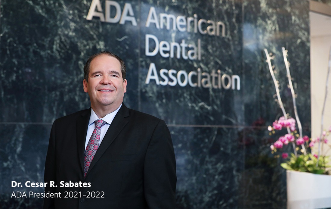 A photograph of the ADA president 2021-2022, Dr. Cesar R. Sabates, standing in the lobby of the ADA building in front of the American Dental Association sign.