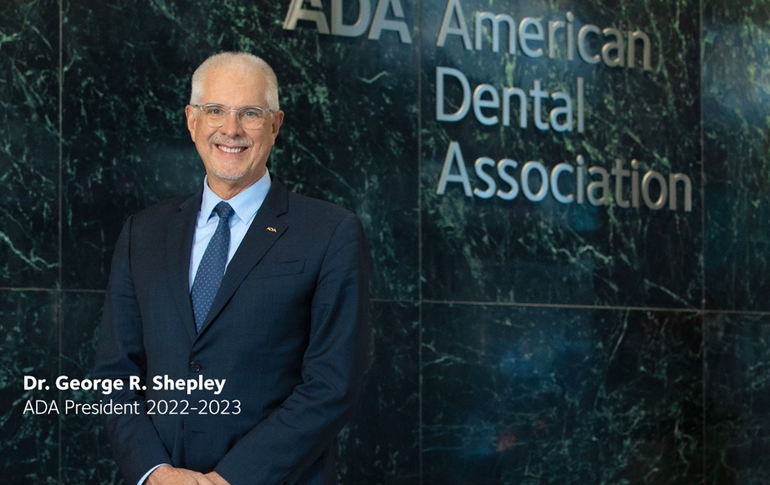 A photograph of the ADA president 2022-2023, Dr. George Shepley, standing in the lobby of the ADA building in front of the American Dental Association sign.