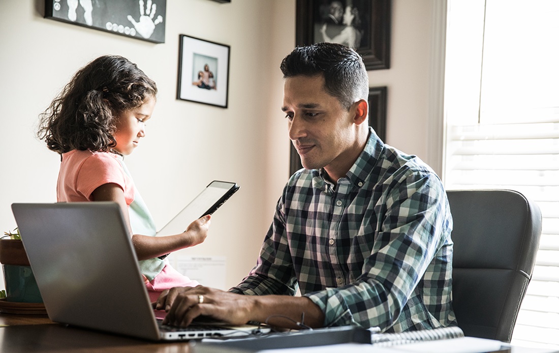 Man in home office working on his laptop with his daughter next to him on a tablet