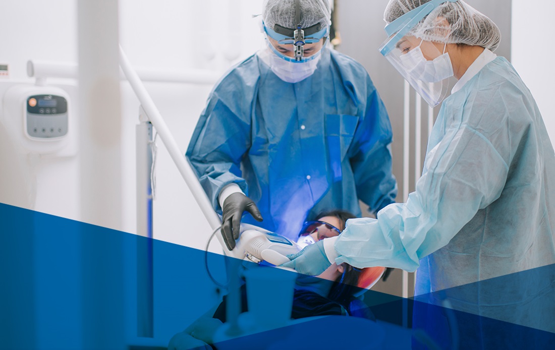 A dentist and assistant treating a patient with face shields and disposible gowns on 