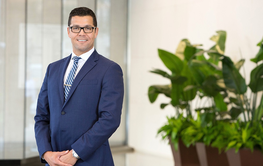 A photograph of Araujo Marcelo Araujo, DDS, MS, PhD standing in a lobby next to three potted plants.