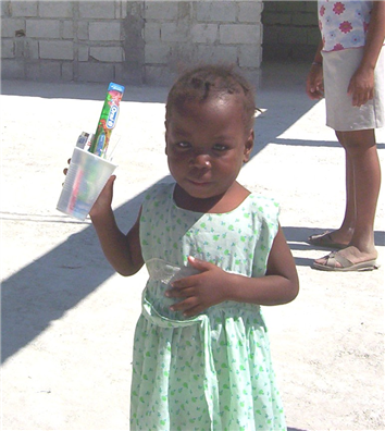 Photo of a child in green dress holding a plastic cup with a tootbrush and toothpaste in it