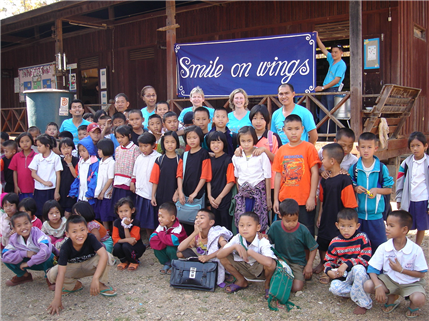 Photo of a group of about forty kids and seven adults posing near a Smiles on wings sign